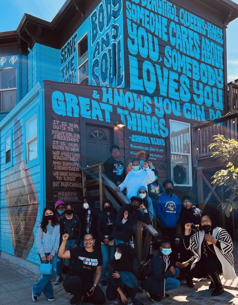 16 Students and teachers of color pose in front of a bright blue building painted with quotes from Sandra Bland and Assata Shakur.