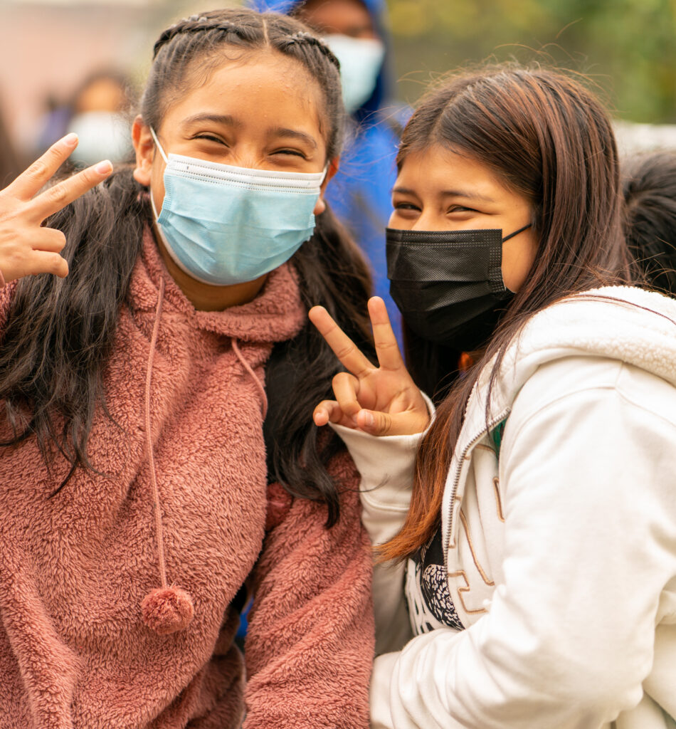 Two students are outside in medical masks throw peace signs and smile at the camera