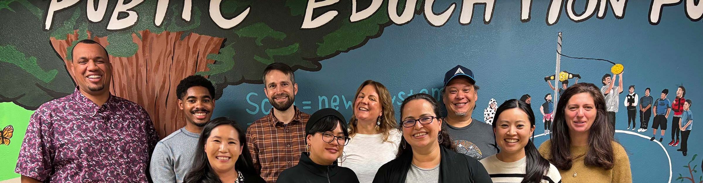 Ten people stand smiling at the viewer in front of a mural of children with "Public Education" in the background.